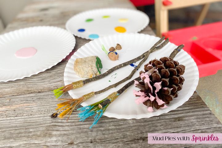 three white paper plates filled with pink, greens, blues and yellow paint and rocks, acorn cups and hickory nut stamps and pine cone glued to a stick for a paint roller