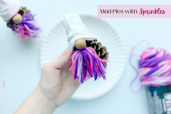 hand holding pine cone gnome with white sock hat, hickory nut nose, and purple and pink yarn beard above a white plate with purple and pink yarn and pine cone gnome sitting on a white table below