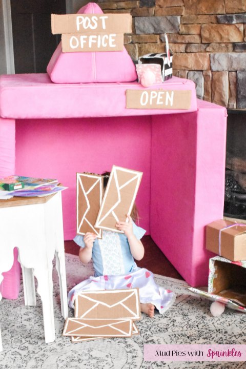 Post office build from a pink play couch (Nugget) with cardboard signage, and a writing center on a white table with a toddler sitting and holding two cardboard envelopes over her face
