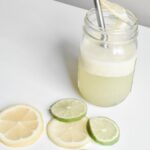 Electrolyte drink for kids in a mason glass jar with stainless steel straw wedged with a lemon on the rim of the glass and slices of lemons and limes spread along a white tabletop