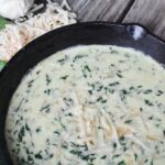 asiago creamed spinach sprinkled with Parmesan cheese in a large black skillet sitting on an old barn wood table with shredded cheese, garlic cloves, and spinach leaves sitting beside the skillet.