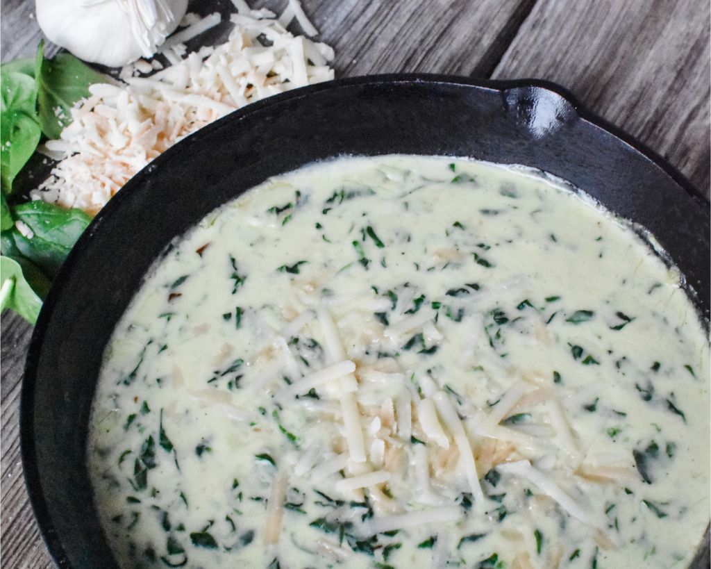 asiago creamed spinach sprinkled with Parmesan cheese in a large black skillet sitting on an old barn wood table with shredded cheese, garlic cloves, and spinach leaves sitting beside the skillet.