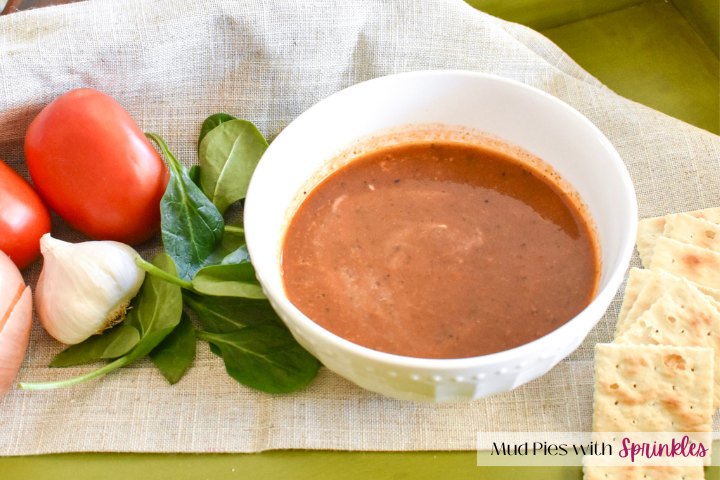 White bowl filled with tomato soup recipe for toddlers with Roma tomatoes, head of garlic, onion and spinach leaves on the left of the bowl and crackers on the right side of the bowl. The bowl of tomato soup is resting on a beige kitchen towel in a green serving tray.