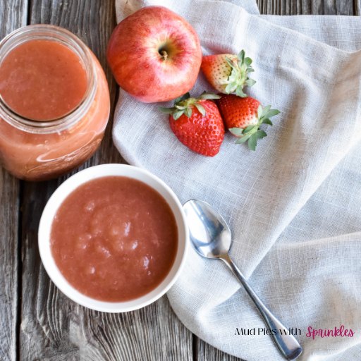 Three Ingredient Applesauce single serving in a white ceramic bowl sitting on a worn wooden table with a beige kitchen towel, silver spoon, three strawberries, a red apple, and a quart jar full of strawberry-applesauce.