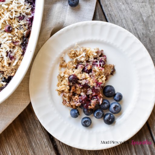 A white baking dish filled with baked oatmeal topped with chocolate chips, shredded coconut, and fresh blueberries rests on a rustic table with a single serving of blueberry coconut baked oatmeal on a white plate.