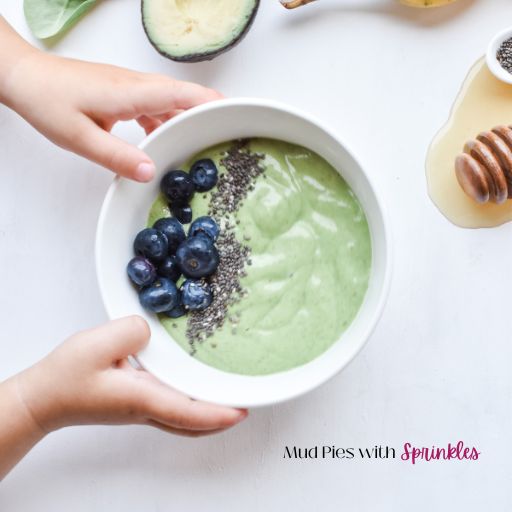 Kid friendly avocado green smoothie recipe in a white bowl topped with blueberries and chia seeds with two little hands grabbing the bowl.