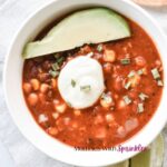 A white ceramic bowl filled with beef enchilada soup recipe topped with green onions and fresh avocado.